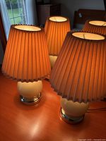 Photo showing 3 small working table lamps with ivory bases and orange-brown pleated shades lit on a wooden surface.