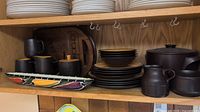 Photo showing a set of Mikasa Terra Stone Paprika dinnerware for four, black mugs, plates, bowls, a black casserole with lid, and a wooden 'crackers' serving tray on a shelf.