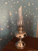 Full view of silver tone oil lamp with glass chimney showing rounded base and classic design against floral wallpaper.
