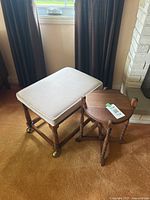 Photo showing a medium wood rectangular ottoman with beige corduroy fabric cushion and small round wooden plant stand with legs and handle, placed on carpeted floor near window and fireplace.