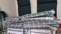 Stack of four folded chair cushions with plaid pattern showing green, navy blue, beige, and white colors, placed on white folding chairs.