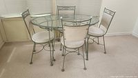 Oval metal framed glass-topped patio table with four matching cushioned metal chairs around it on carpeted floor in a room.