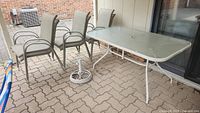 Rectangular patio table, metal frame and textured tempered glass top, with six beige sling style stacking chairs and metal umbrella base on patio surface.