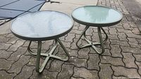 Two round patio side tables placed outdoors on brick pavers showing metal frames and glass tops