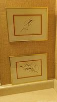 Two framed Benjamin Chee Chee prints displayed vertically on a wall, showing simple line art with orange mats and thin gold frames.