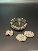 Sterling silver engraved bangle bracelet, two oval brooches, and clip-on earrings shown from above on a dark surface.