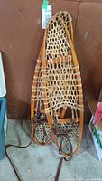 Pair of traditional rawhide snowshoes leaning against wall, showing full length with leather straps and wooden frames visible.
