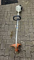 Full length view of Stihl gas powered string trimmer with orange cutting guard and white engine casing lying on paved surface.