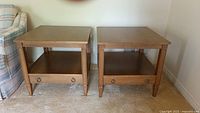 Pair of matching wood end tables side by side against a wall on beige carpet, showing full tables with storage drawers and brass ring pulls.