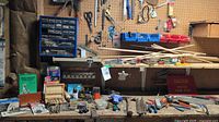 Wide view of workbench showing tools, wooden dowels, pliers, files, drill bits in cases, and pegboard with additional tools and small hardware drawers