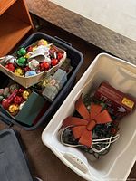 Two plastic storage bins on the floor near a bed; one black bin filled with assorted Christmas ball ornaments, another white bin with Christmas lights and a red bow.