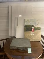 Full view of six canvases with varying sizes and one painted canvas leaning on a table, alongside a closed vintage metal brush storage box with label 'Lot 60'.