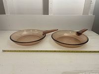 Two 10 inch diameter amber tempered glass frying pans with long handles, sitting side by side on white surface with measuring tape in front showing size.