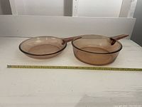 Two Corning Visions cooking pans on white surface with measuring tape showing 10-inch diameter and approximately 2-inch and 3-inch depths