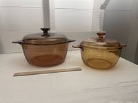 Two amber glass Corning Visions pots with lids side-by-side with ruler in front showing size.