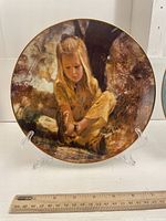 Front view of the decorative plate showing detailed painted image of young girl with sand in shoe, displayed on clear stand with ruler for scale.