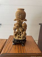 Front view of the tan soapstone Foo Dog carving with lattice vase and dark wooden base on wooden table against white panel background.