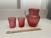Photo showing the full set: one cranberry glass pitcher and two matching cranberry glass cups with ruler for scale.