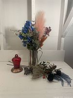 Photo showing assorted artificial flowers and foliage arranged with the stainless steel pitcher and hummingbird feeder on a white surface against a white wall.
