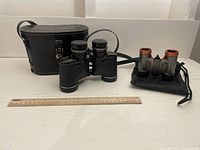 Wide shot showing two binoculars; Tasco binoculars with black textured body and leather carrying case, and smaller Pentax binoculars with orange/red eye cups on a soft black pouch, ruler for scale.