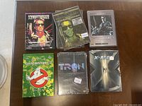 The seven DVDs laid out on a wooden surface showing front covers: The Terminator (three versions), Terminator 2, Ghostbusters 1 & 2 set, Tron 20th Anniversary edition, and X-Men special edition.