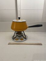 Side view of vintage yellow enamel fondue pot set on metal stand with bottom plate, ruler in front for scale.