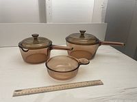Three amber glass cookware pieces including two pots with lids and one grab-it bowl with a handle, photographed beside a ruler for scale.