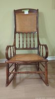 Front view of the antique wooden rocking chair shows cane back and seat with spindle detailing and curved rockers.