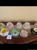 Display of nine assorted fine bone china teacups and saucers on a wooden table against a plain wall.