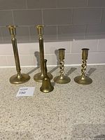 Full view of two tall smooth brass candlesticks, two short twisted brass candlesticks and one small brass bell on countertop.