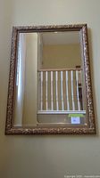Full view of the large gold-colored framed mirror hanging on a wall reflecting stair railings and a neutral wall.