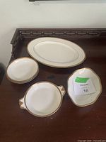 Top-down view of seven white porcelain serving dishes with gold rims including serving platter, bowls with handles, and small bowls arranged on wooden surface