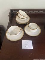 Photo showing 3 white porcelain teacups stacked inside each other and 4 white saucers with gold rims, placed on a wooden surface.