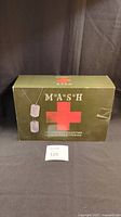 Closed green military-themed M*A*S*H box with red cross and dog tag graphics, showing front of the collection box.