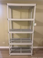 Full view of white plastic shelving unit with five shelves and black corner accents, standing against a wall.