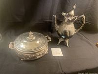 Full view of silver casserole dish with lid and matching silver tea pot on black background.