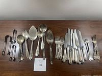 Various silver and stainless steel serving utensils laid out on a wooden table in one row. Includes forks, spoons, serving spoons, tongs, and knives of assorted patterns.
