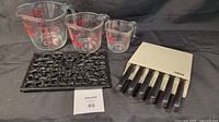 Lot items displayed including three glass measuring cups with red markings, black decorative trivet, and Wiltshire cutting utensil set in white block with black handles.