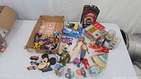 Box containing mixed vintage toys including Thomas the Train 2005 with track, Mickey Mouse radio missing battery cover, Sesame Street cake decor, and various character figurines.