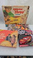 Photo of four board games: Mark "three" vintage game, Dwight D. board game, Cars Lightning McQueen game, and Jurassic World Volcano game arranged on a table.