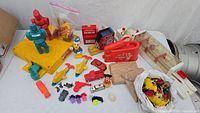 Full assortment of vintage plastic and wooden toys spread on a white surface showing the Rock 'Em Sock 'Em Robots game, various small toys including a wooden shoe tying toy, plastic figurines, and a bag of small animals.