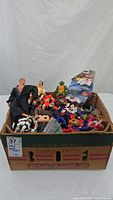 Box filled with mixed action figures and toys including large humanoid figures and smaller colorful character figurines.