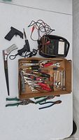 Full view of the assortment of hand tools including pliers, wrenches, screwdrivers, an axe, two black caulking guns, and the Simpson electrical tester.