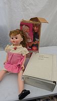 Photo showing Vintage Little Miss Echo doll seated with legs spread, wearing a pink dress with lace collar, black shoes, original box and white box labeled 'Porcelain Tee Head' nearby