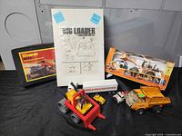 Vintage construction toys lot arranged on a black surface showing boxed Stomper carrying case, Big Loader 1 Construction Set instruction manual, small construction trucks, and Transcon truck toy.