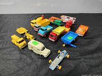 Overview of approximately 12 various die cast toy trucks including yellow construction vehicles, blue and red delivery trucks, ambulance, utility trucks, and trailers arranged on black fabric