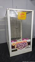 Pretzel warmer display case front view showing the glass panels, white metal frame, promotional stickers, and base