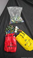 Collection of marbles spilling out of two labeled fabric bags and one plastic bag, displayed on black background