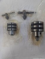 Two drill press chucks and their keys wrapped in plastic on a white surface.