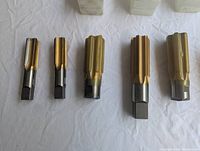 Five new machinist taps laid out in a row showing their spiral flutes and squared shanks from the side.